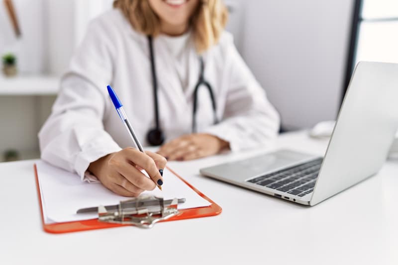 Physician completing a medical record beside a laptop